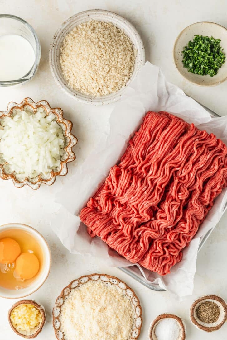 White bowls of onion, breadcrumbs, parmesan, eggs, spices, and milk next to ground beef on a white counter.
