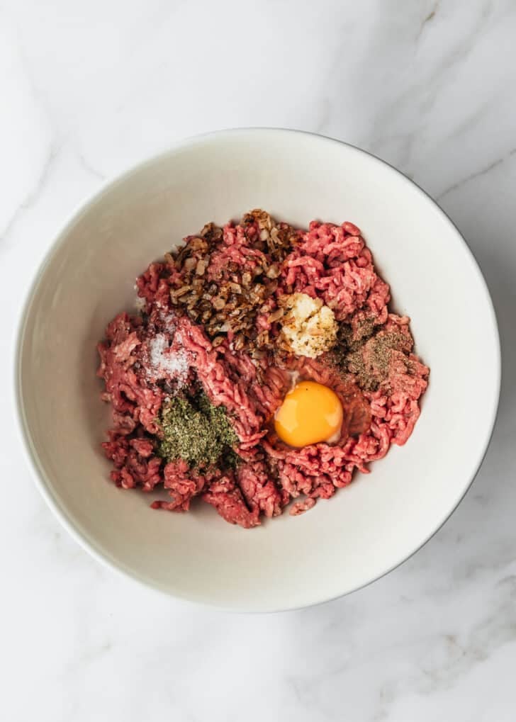 A white bowl of ground beef, spices, and an egg on a white table.