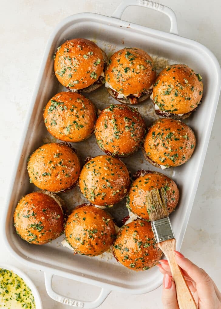 A hand using a pastry brush to brush a white pan of Italian meatball sliders with garlic butter.