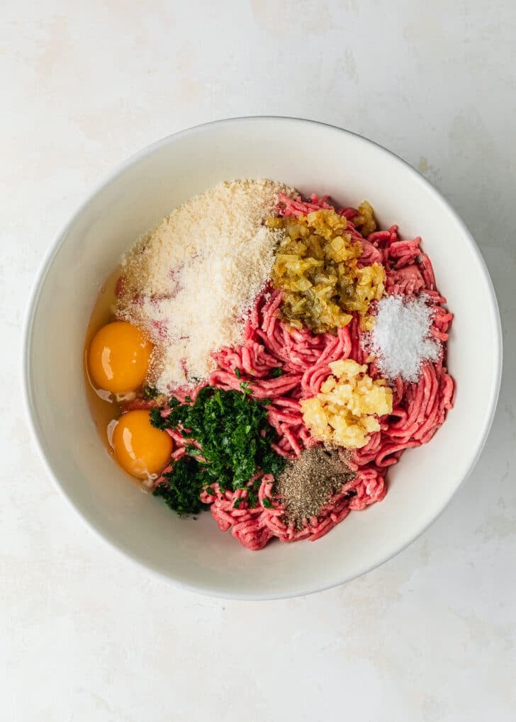 A white bowl of ground beef, parmesan, eggs, and spices on a white counter.