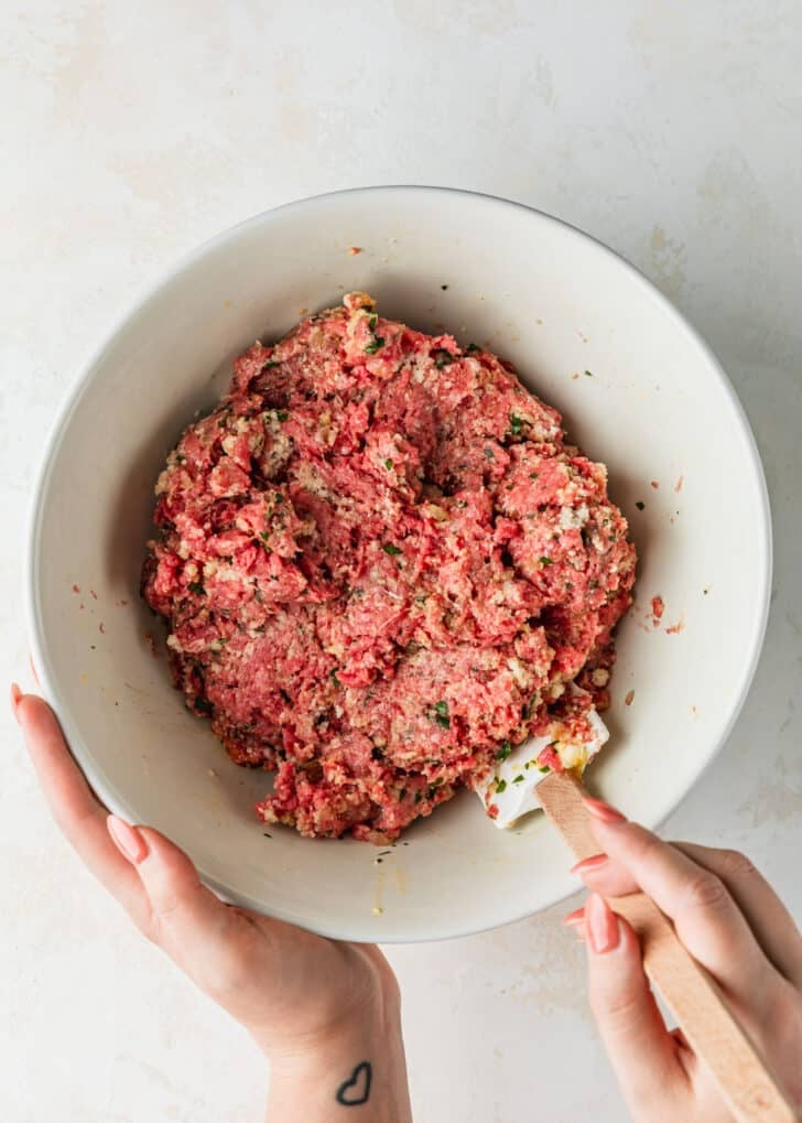 Hands using a rubber spatula to mix meat in a white bowl.