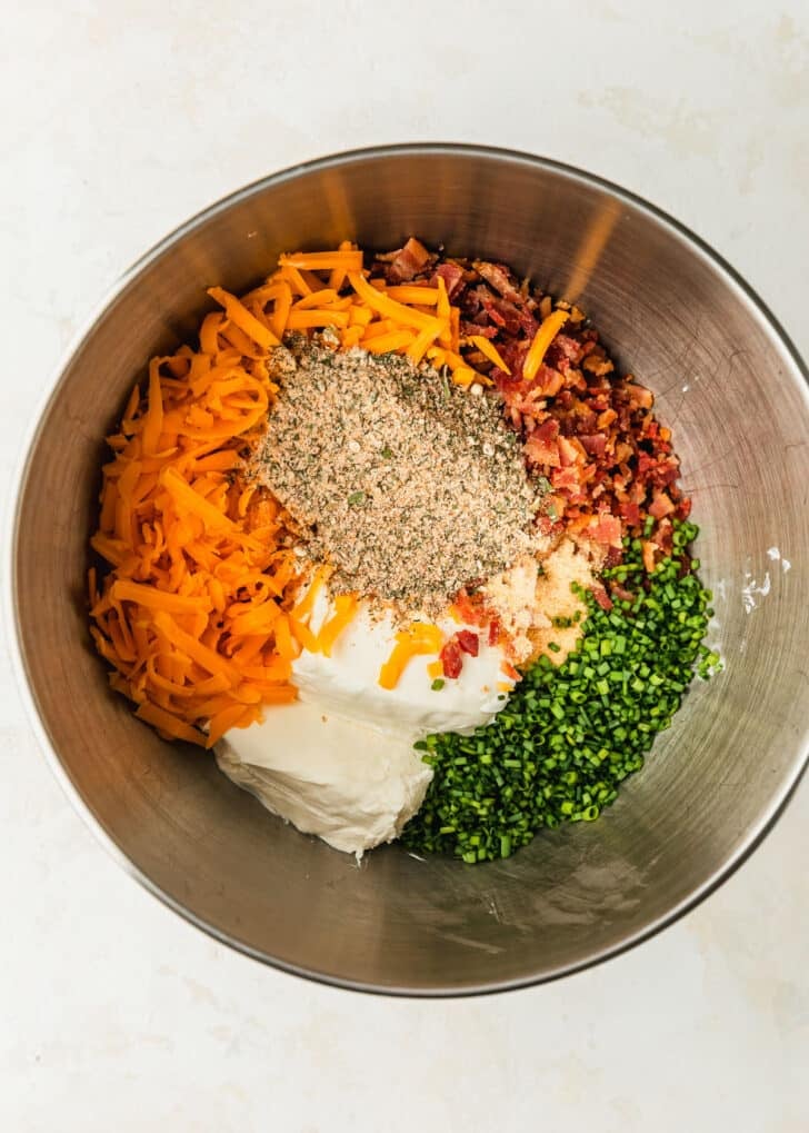 A silver bowl of cream cheese, cheddar, ranch seasoning, bacon, and chives on a beige counter.