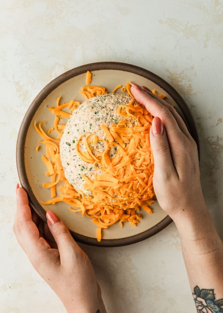 Hands pressing cheddar cheese to a cheese ball on a tan plate.
