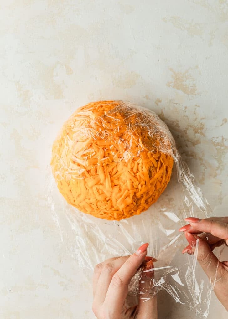 Hands wrapping a pumpkin shaped cheese ball in plastic wrap on a tan counter.