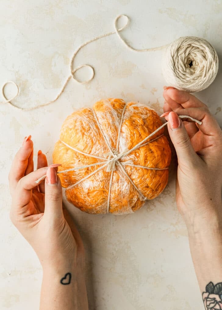 Hands tying string around a pumpkin cheese ball on a tan counter.