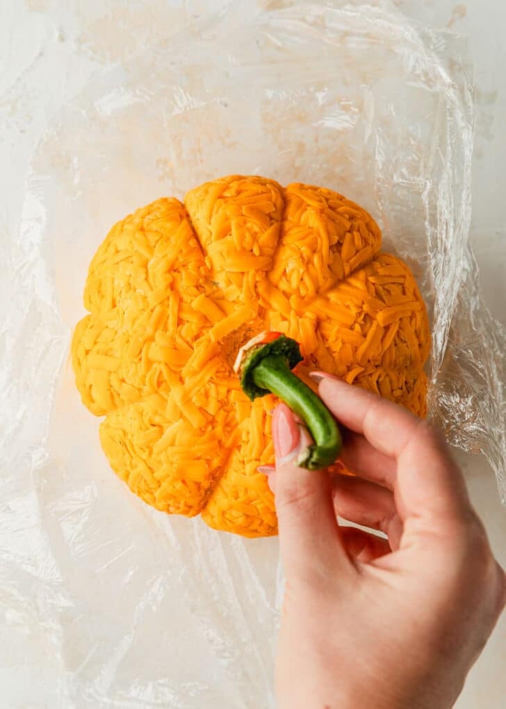 A hand placing a pepper stem on a pumpkin shaped cheese ball.