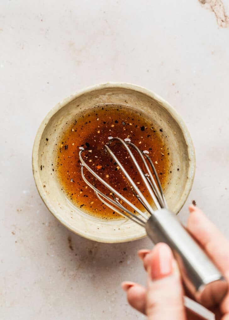 A hand whisking vinaigrette in a white bowl on a tan counter.