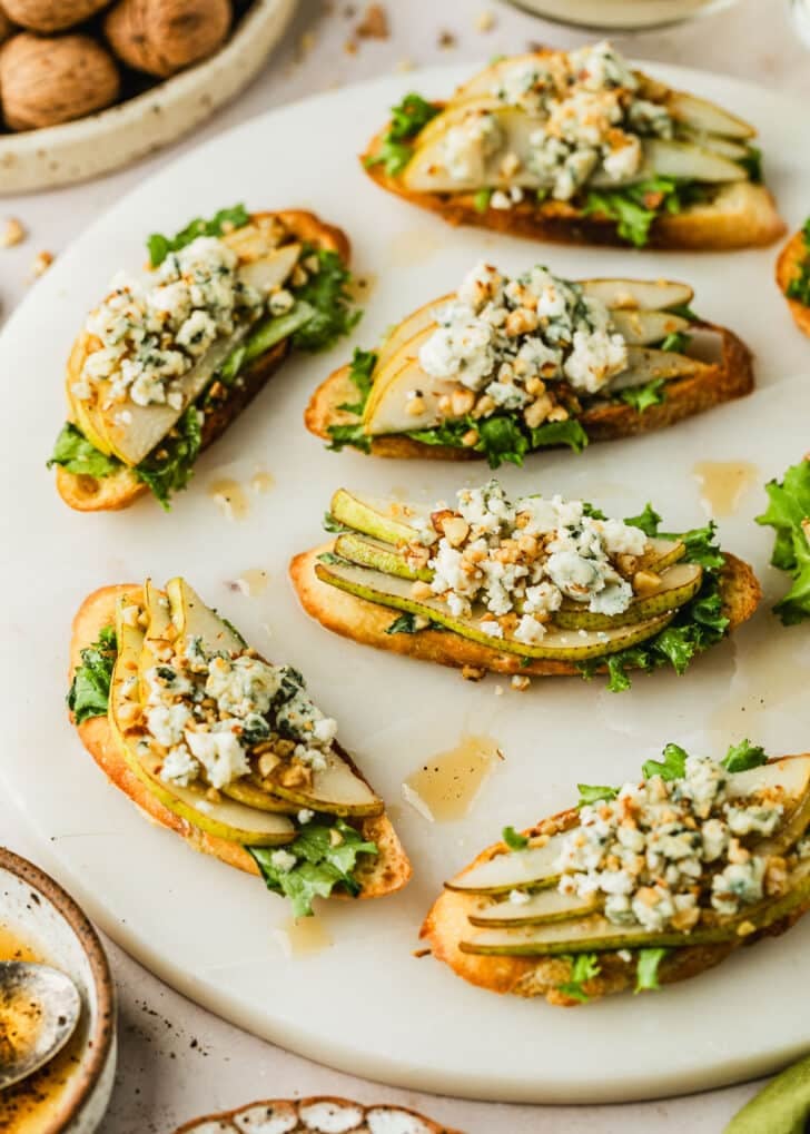 A white platter of pear and blue cheese crostini on a tan counter next to glasses of white wine, a white bowl of walnuts, and a white bowl of vinaigrette.