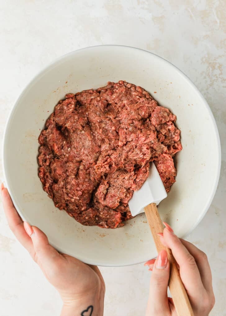 Hands using a rubber spatula to mix ground beef and spices in a white bowl.