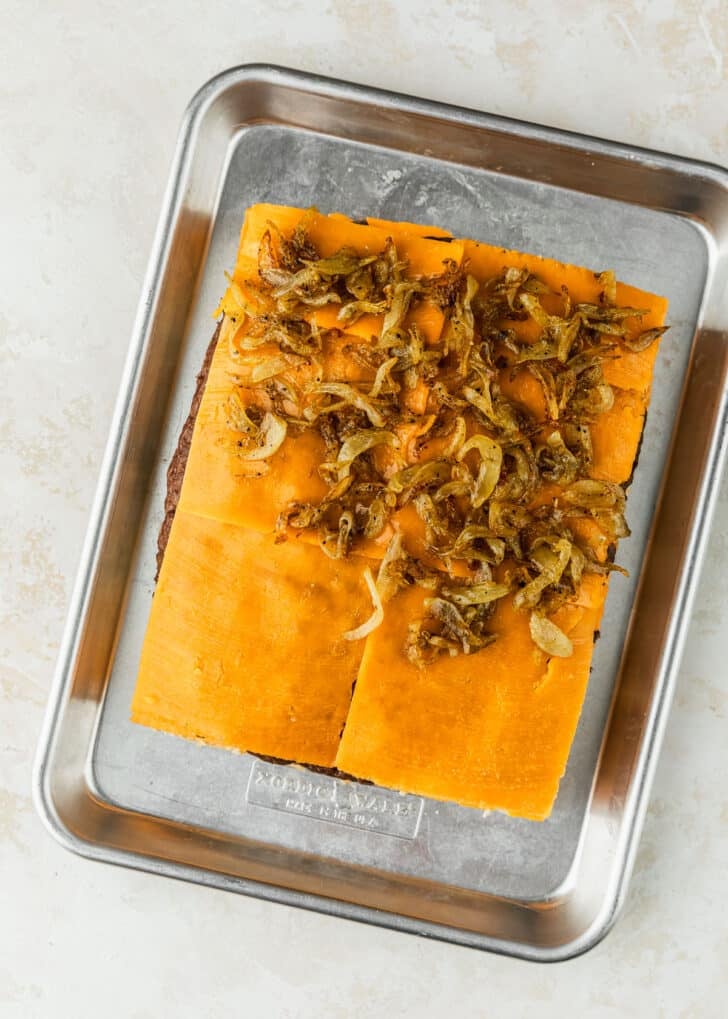 A sheet pan of unbaked cheeseburger sliders with Hawaiian rolls and onions on a tan counter.