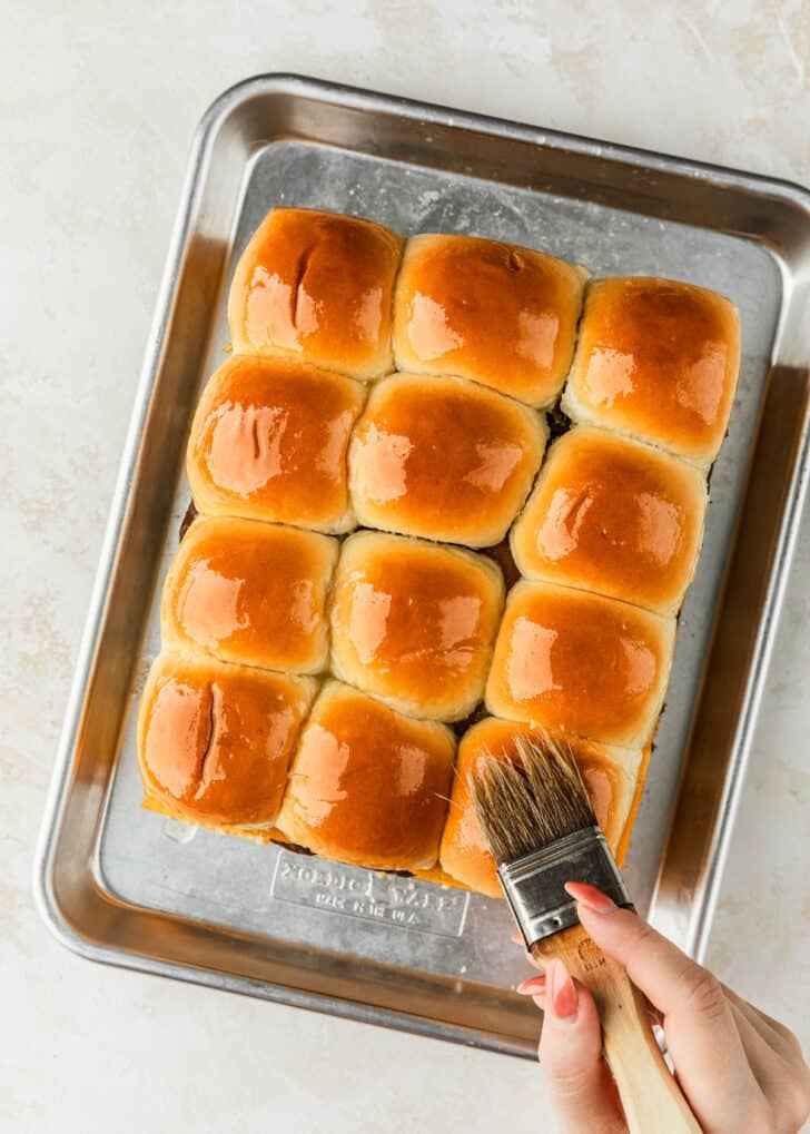 A hand using a pastry brush to brush unbaked cheeseburger sliders on Hawaiian rolls with butter.