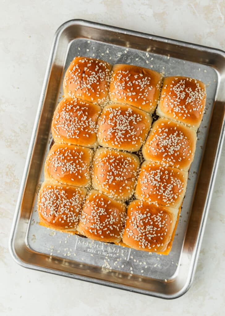 A sheet pan of unbaked Hawaiian roll cheeseburger sliders with sesame seeds on a tan counter.