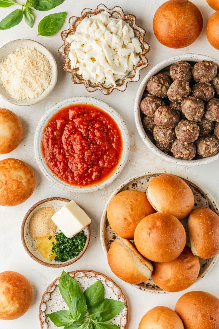 White and brown bowls of rolls, meatballs, red sauce, basil, cheese, and spices on a white counter.