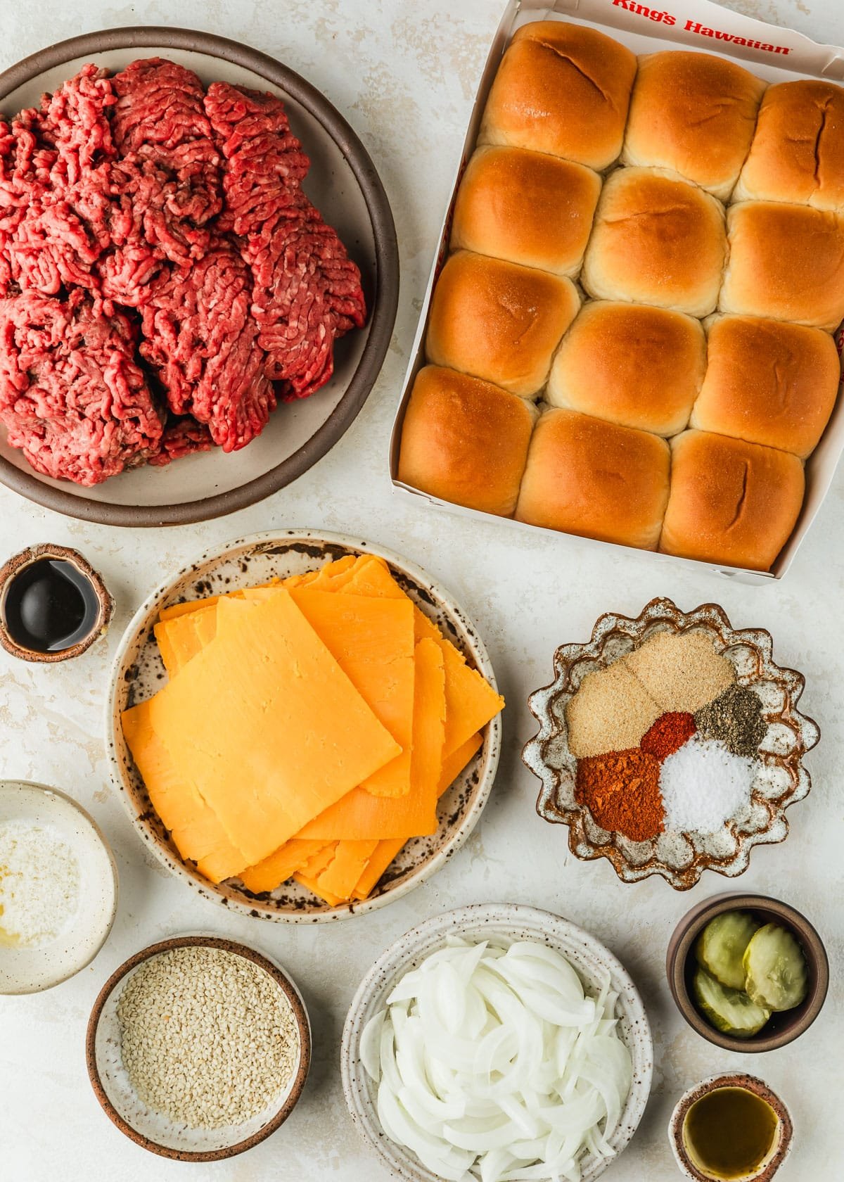 White and brown bowls of ground beef, cheese, Worcestershire sauce, spices, pickles, onions, sesame seeds, butter, and oil on a tan counter next to rolls.