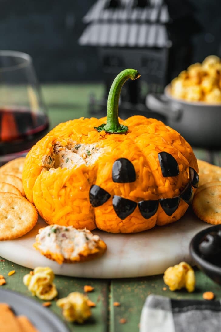 A Halloween pumpkin cheese ball on a white marble tray next to glasses of red wine, a checkered linen, and black bowls of popcorn, crackers, and olives with a green wood background.