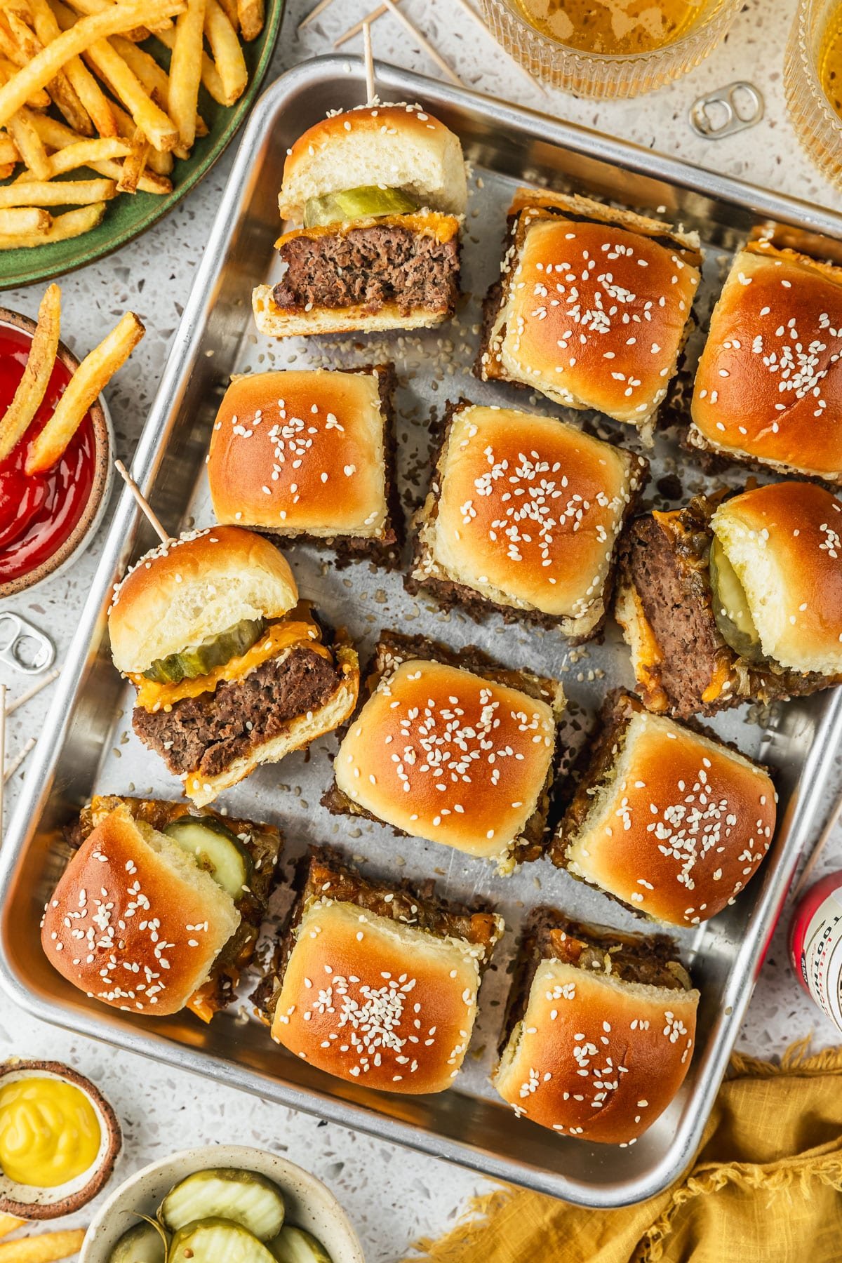 A sheet pan of cheeseburger sliders with Hawaiian rolls on a white counter next to a green plate of fries, glasses of beer, a yellow linen, toothpicks, and brown bowls of ketchup, mustard, and pickles.