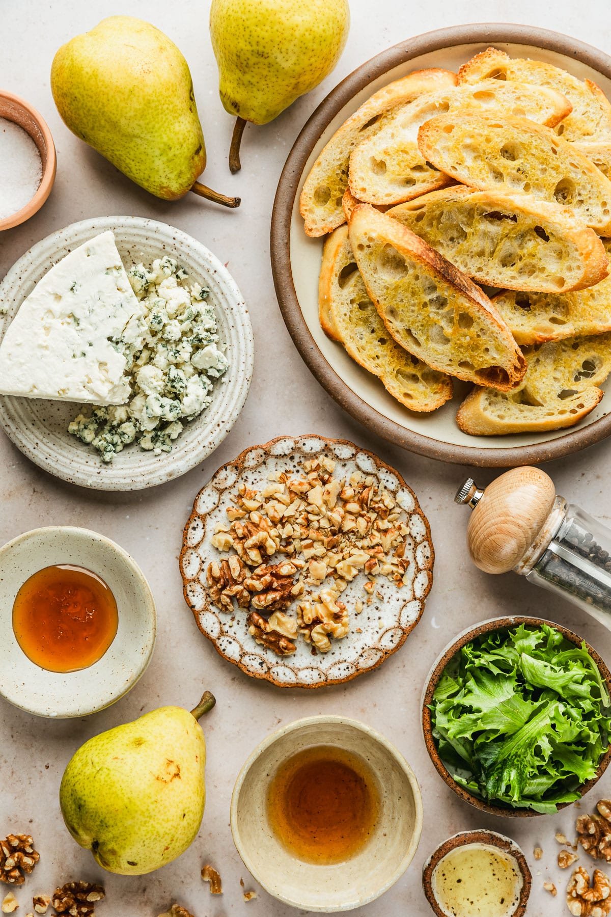 White and brown bowls of bread, cheese, walnuts, greens, spices, honey, and vinegar on a tan marble counter next to pears.