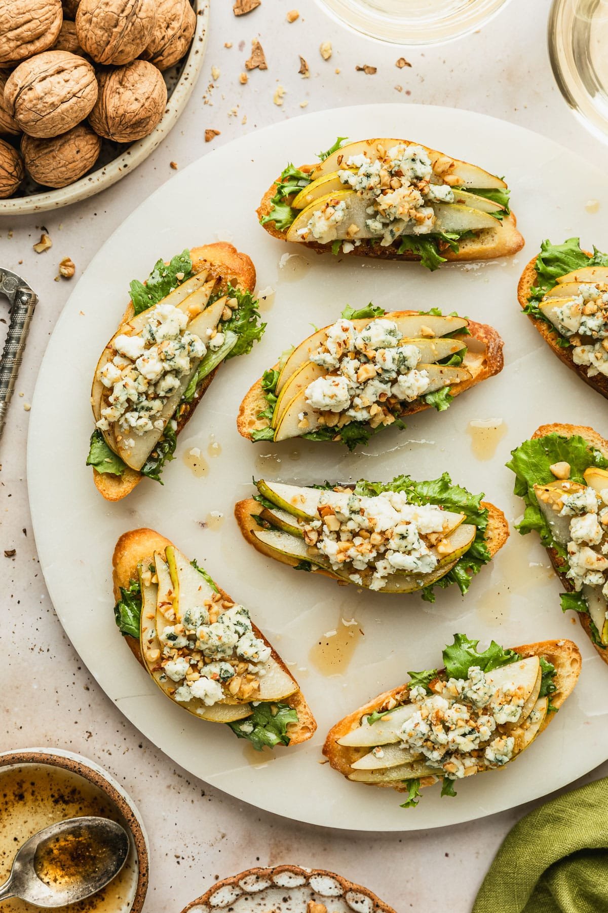 A white platter of pear and blue cheese crostini with honey vinaigrette and walnuts on a tan marble counter next to glasses of white wine, an olive green linen, and a white bowl of walnuts.