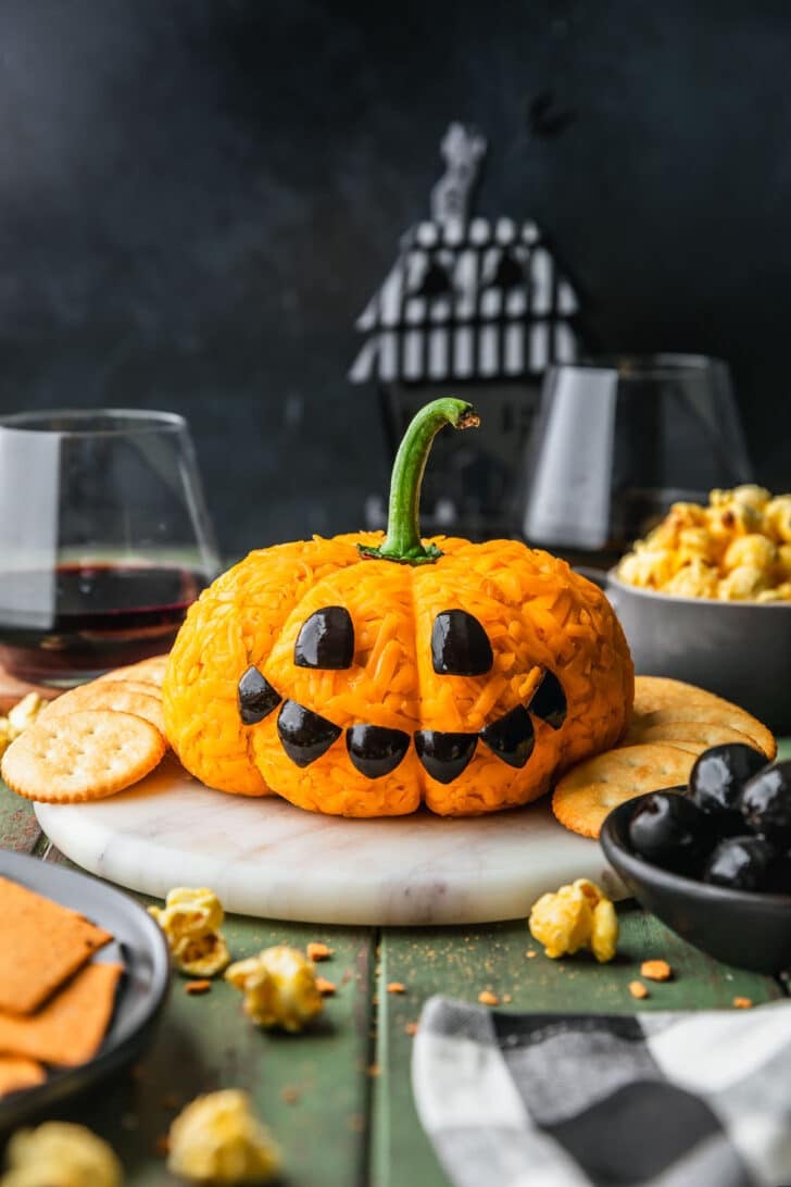 A jack o'lantern cheese ball on a white tray next to black bowls of popcorn, olives, and crackers, glasses of red wine, and a checkered linen on a green counter.