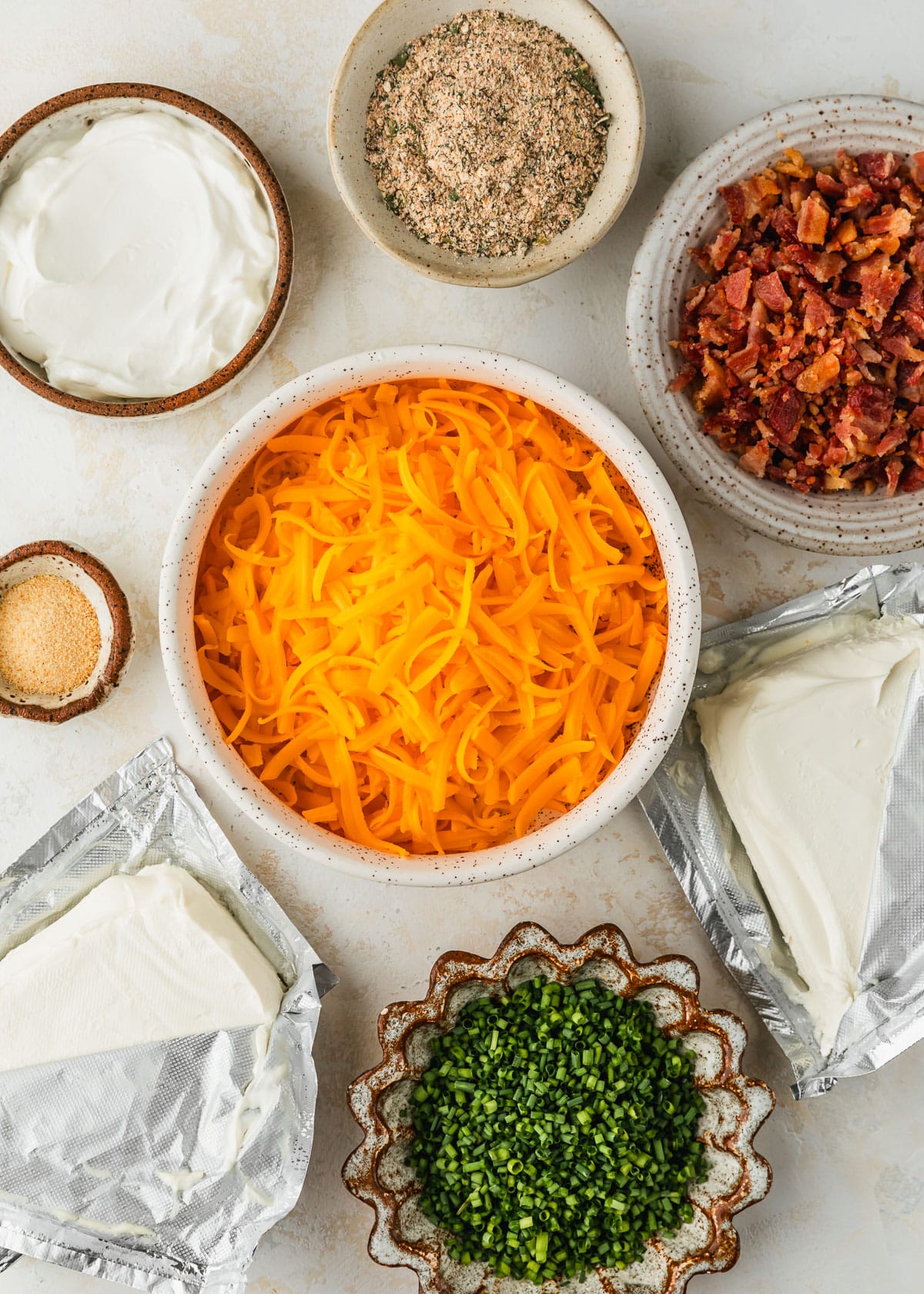 White and brown bowls of cheddar cheese, bacon, ranch seasoning, sour cream, garlic powder, and chives next to bricks of cream cheese on a tan counter.
