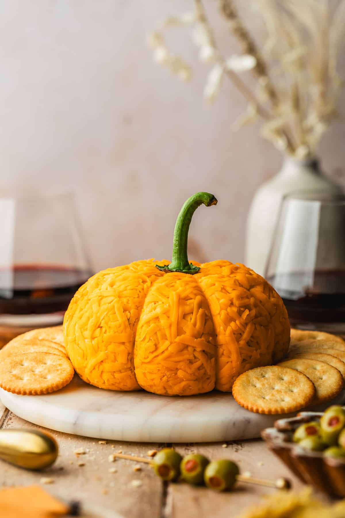 A white marble platter with a pumpkin cheese ball on a wood board next to glasses of red wine, a vase of wheat, and a brown bowl of green olives.