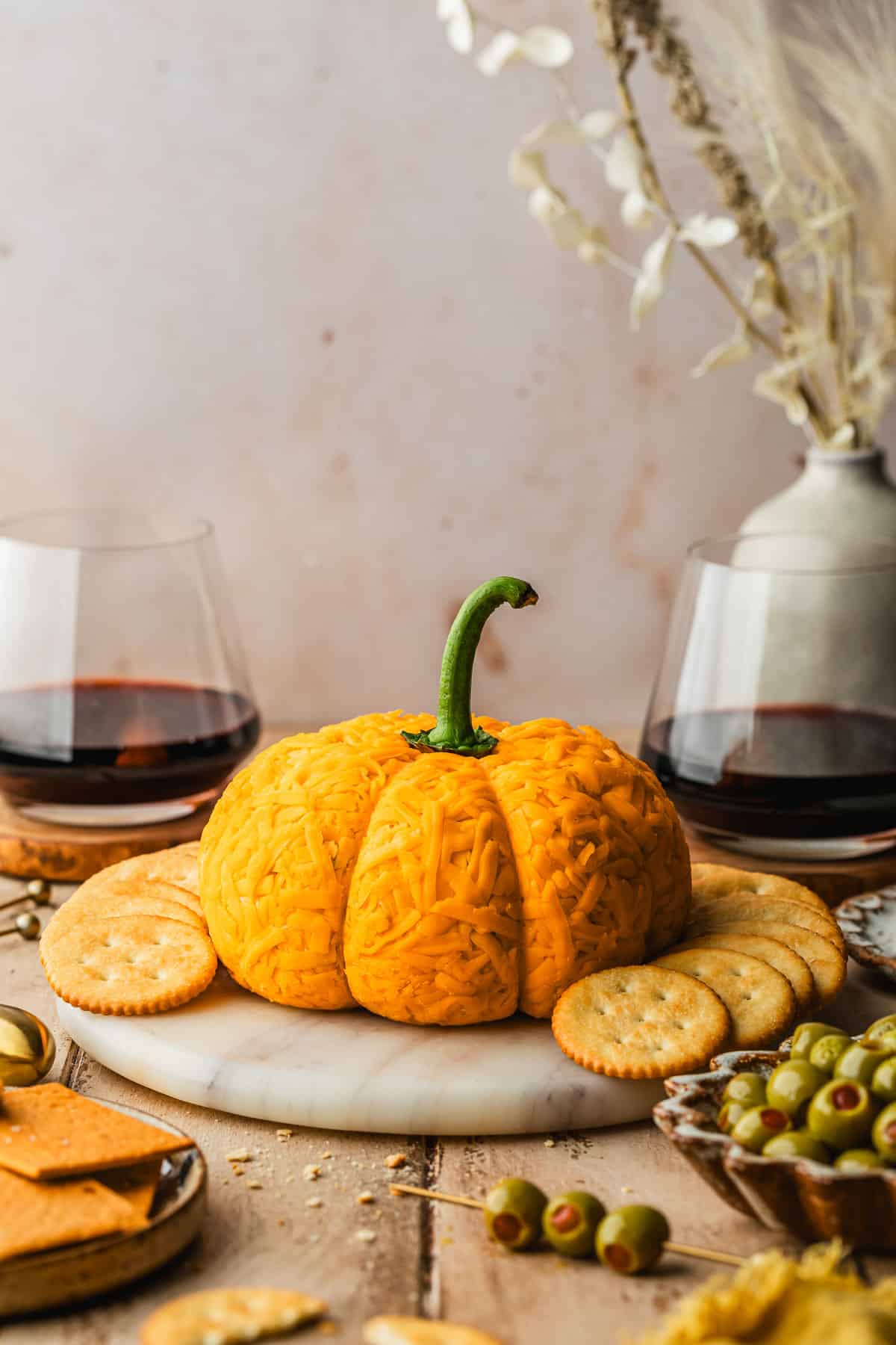 A pumpkin shaped cheese ball on a white board with crackers next to glasses of red wine, a yellow linen, and brown bowls of olives and crackers with a wood background.