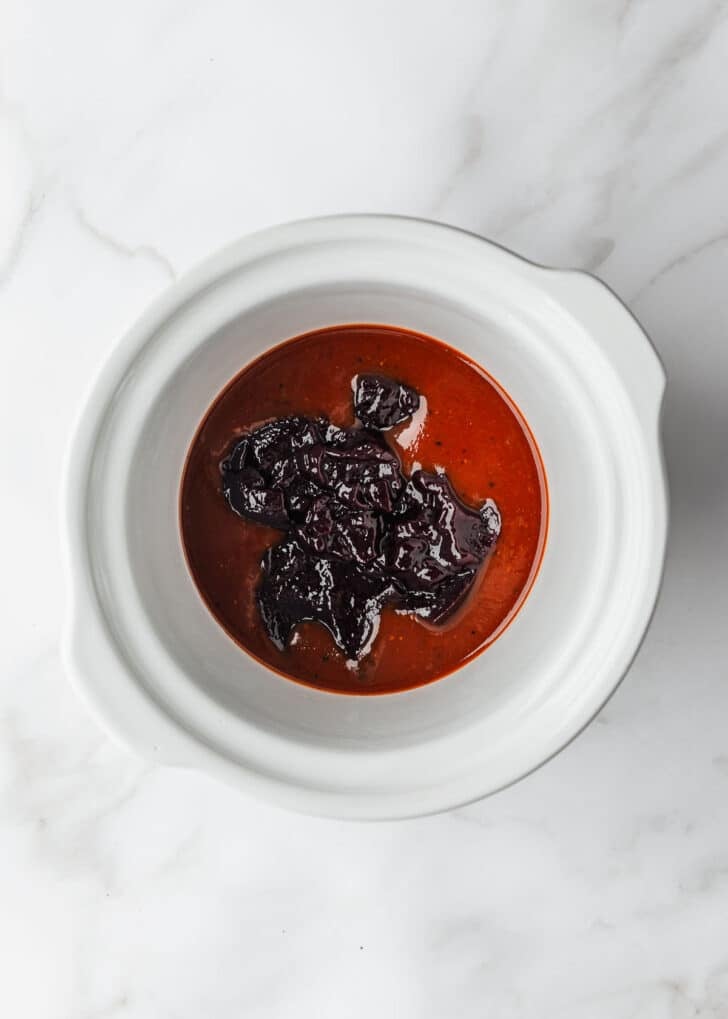 A white slow cooker with BBQ sauce and grape jelly on a white counter.