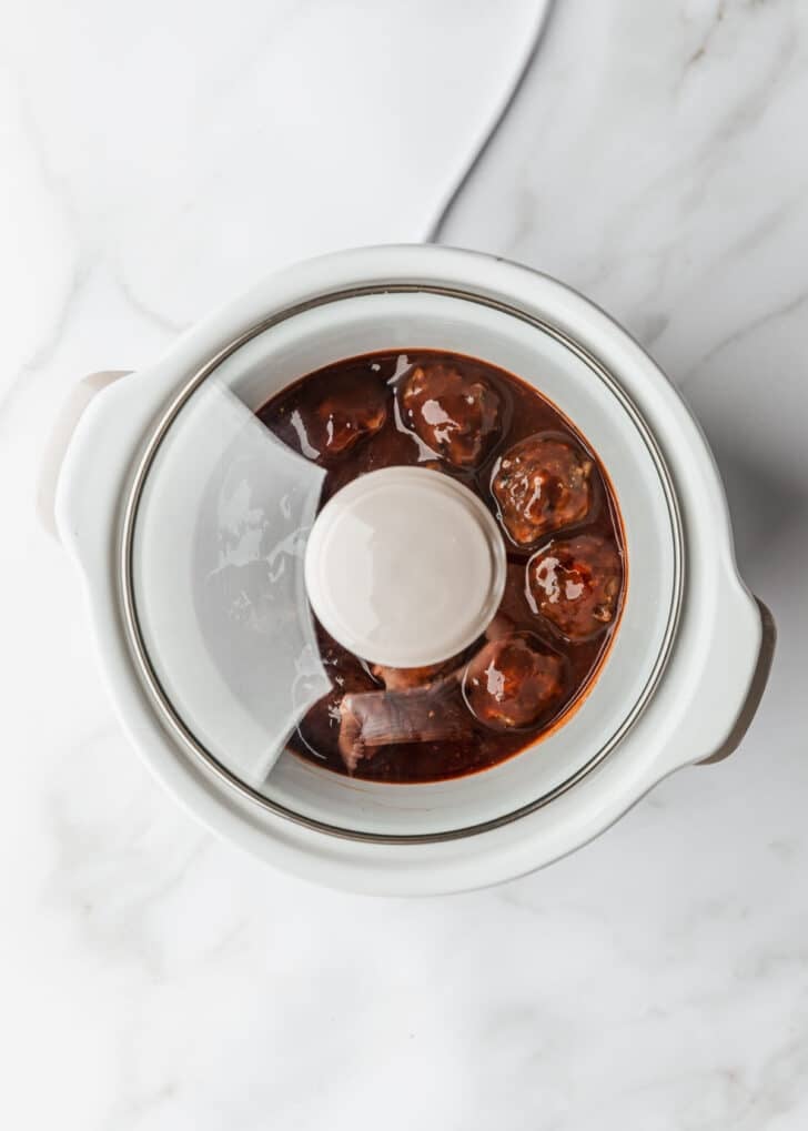 A crock pot of cooked BBQ grape jelly meatballs with a lid on a white counter.