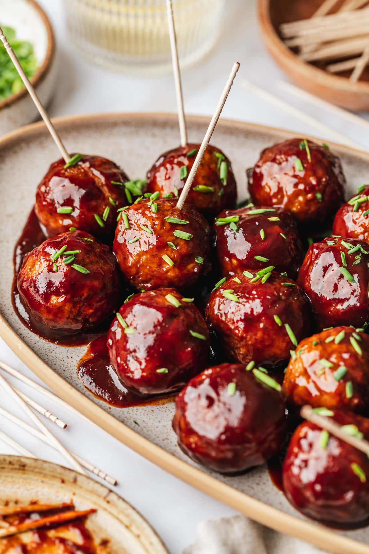 A stoneware platter of BBQ grape jelly meatballs on a white marble counter next to a glass of white wine, wood bowl of toothpicks, and white bowl of chives.