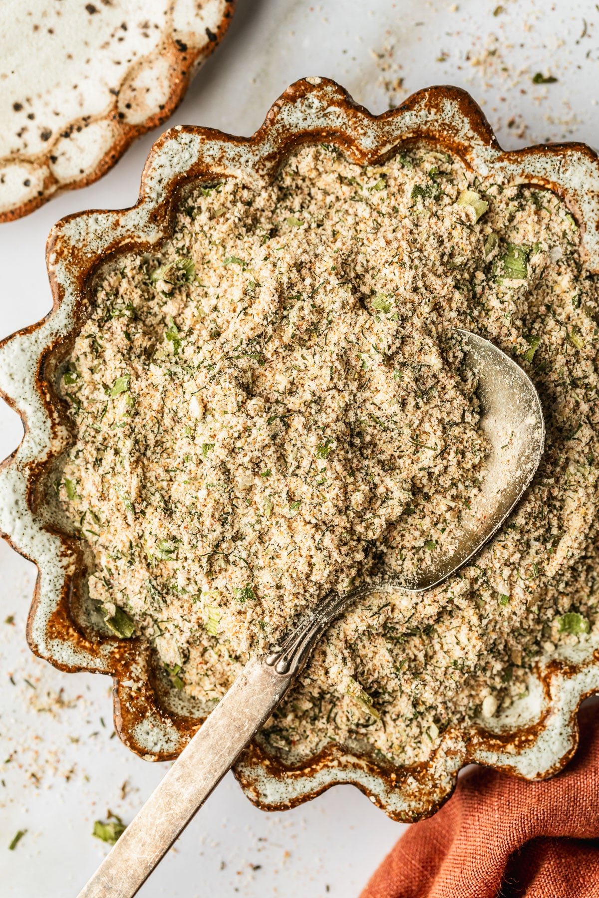 A brown and white bowl of homemade spicy ranch seasoning with a spoon on a white counter next to a white and brown plate and orange linen.