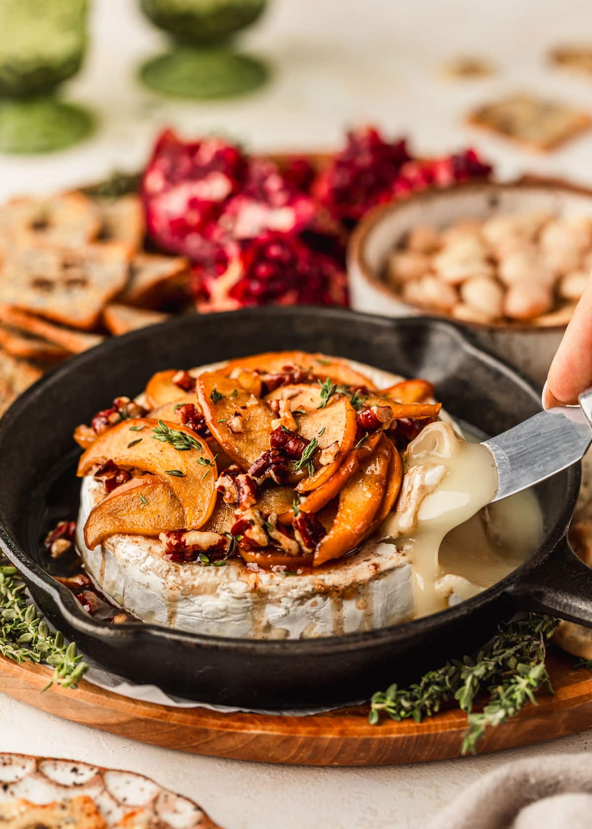 A knife slicing baked brie with apples and pecans in a cast iron skillet. The skillet is on a wood board next to crackers, pomegranates, and a white bowl of almonds.