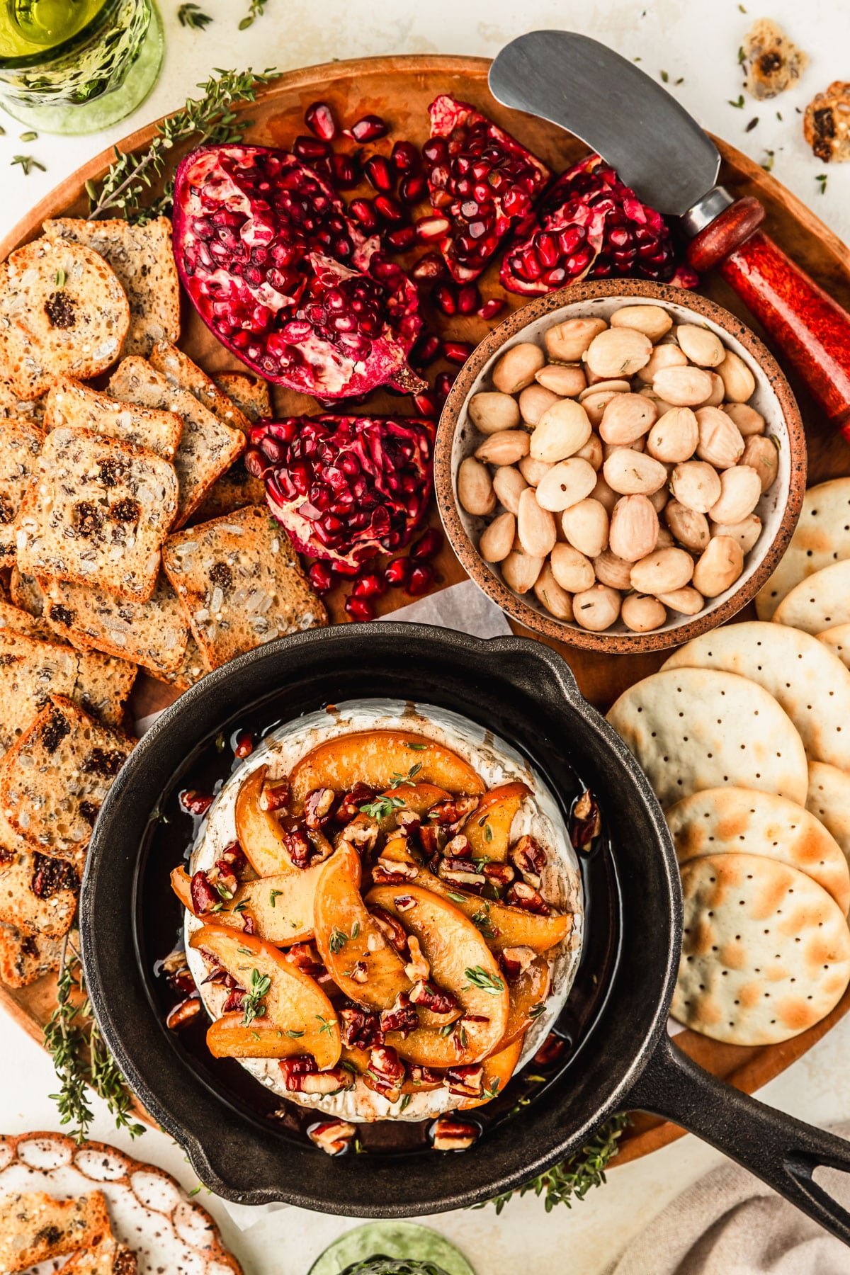 A cast iron skillet with apple baked brie on a wood board next to crackers, pomegranates, and a brown bowl of almonds. The platter is on a beige counter next to green glasses of wine and a beige linen.