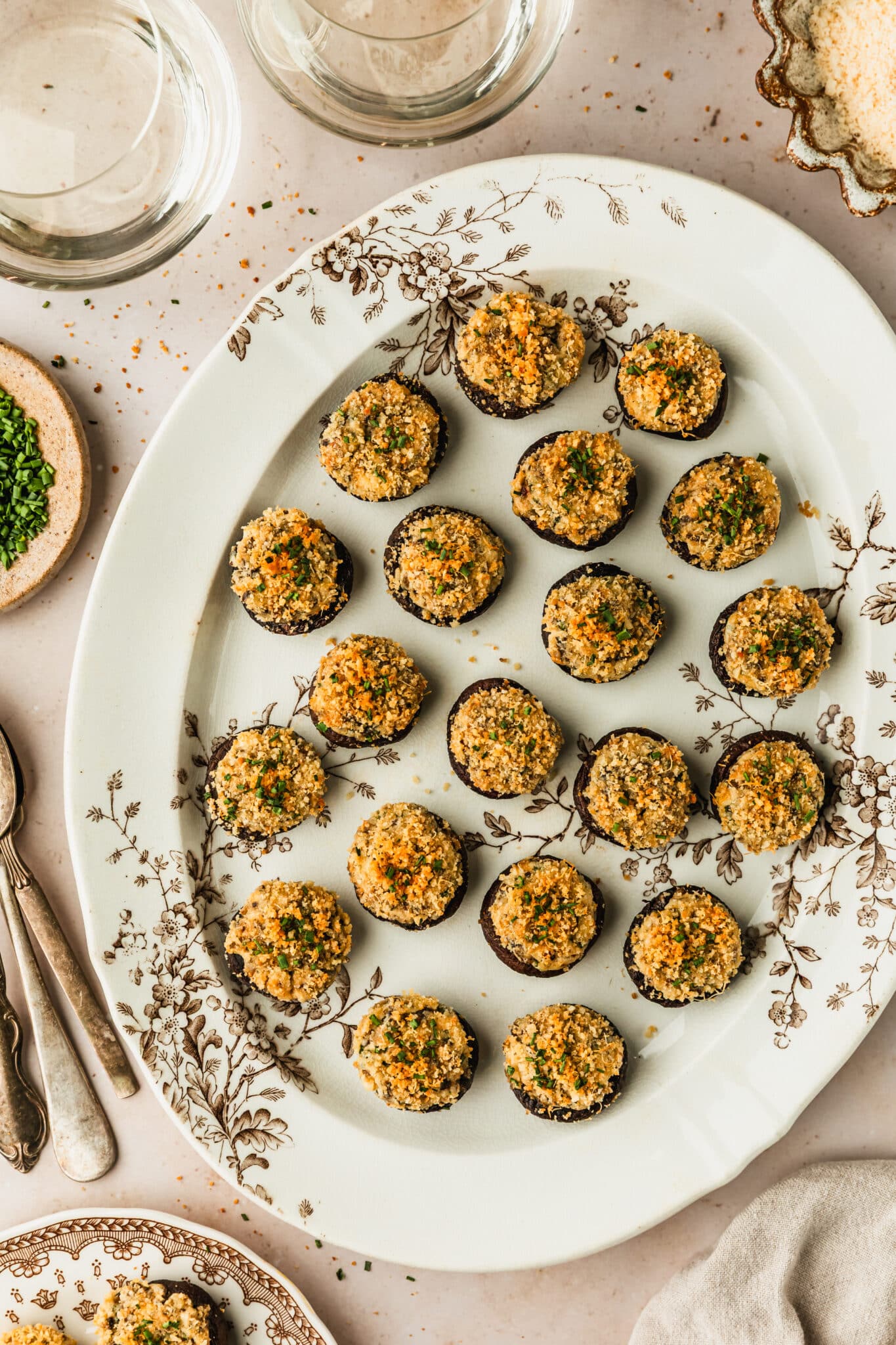 A white and brown floral plate of boursin cheese stuffed mushrooms next to glasses of white wine, a beige linen, and brown plates of parmesan and chives with a tan marble background.