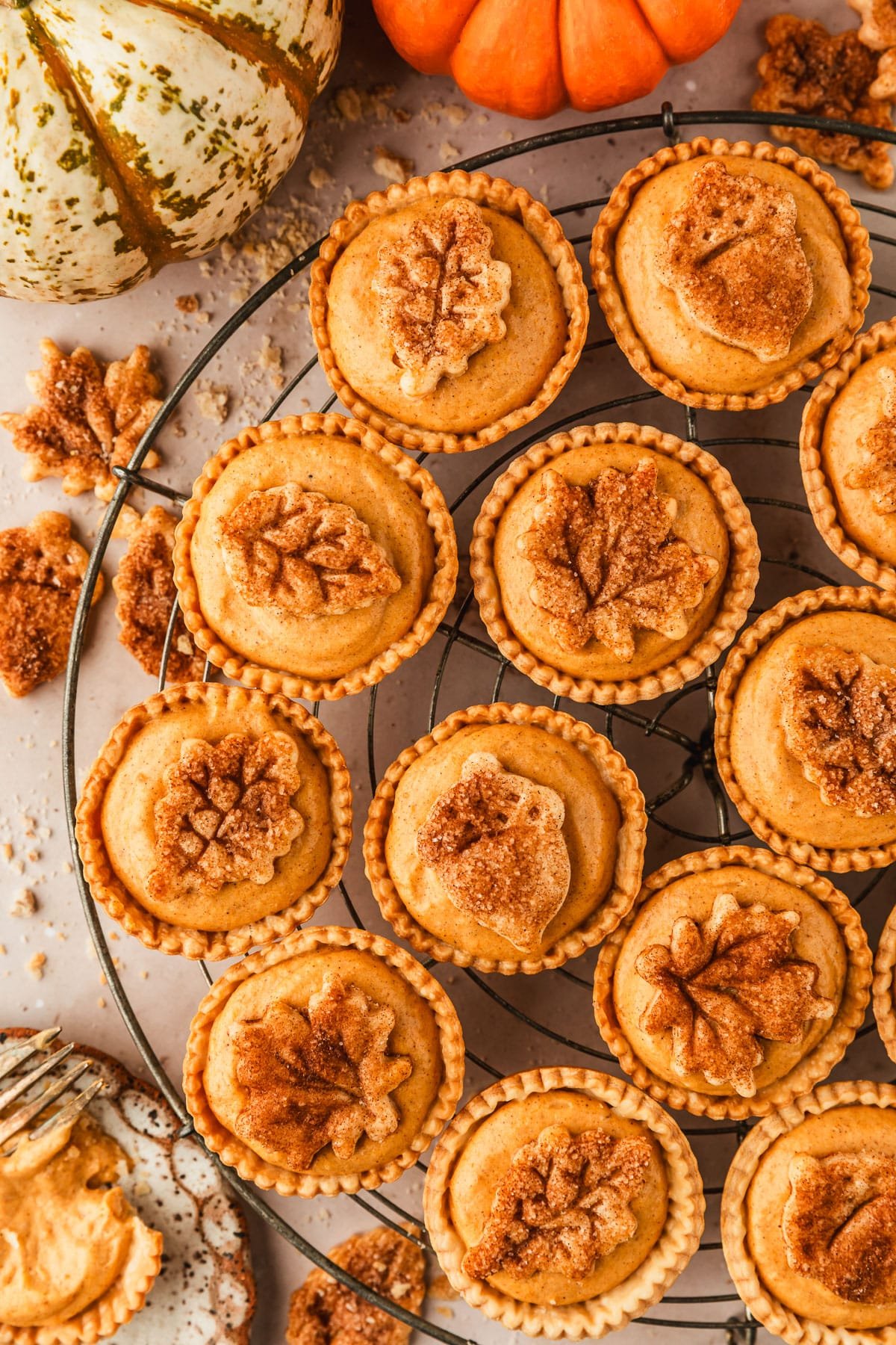 A wire rack of mini pumpkin cream cheese pies topped with pie crust leaves on a beige marble counter next to a white and brown plates, pie crust leaves, and pumpkins.