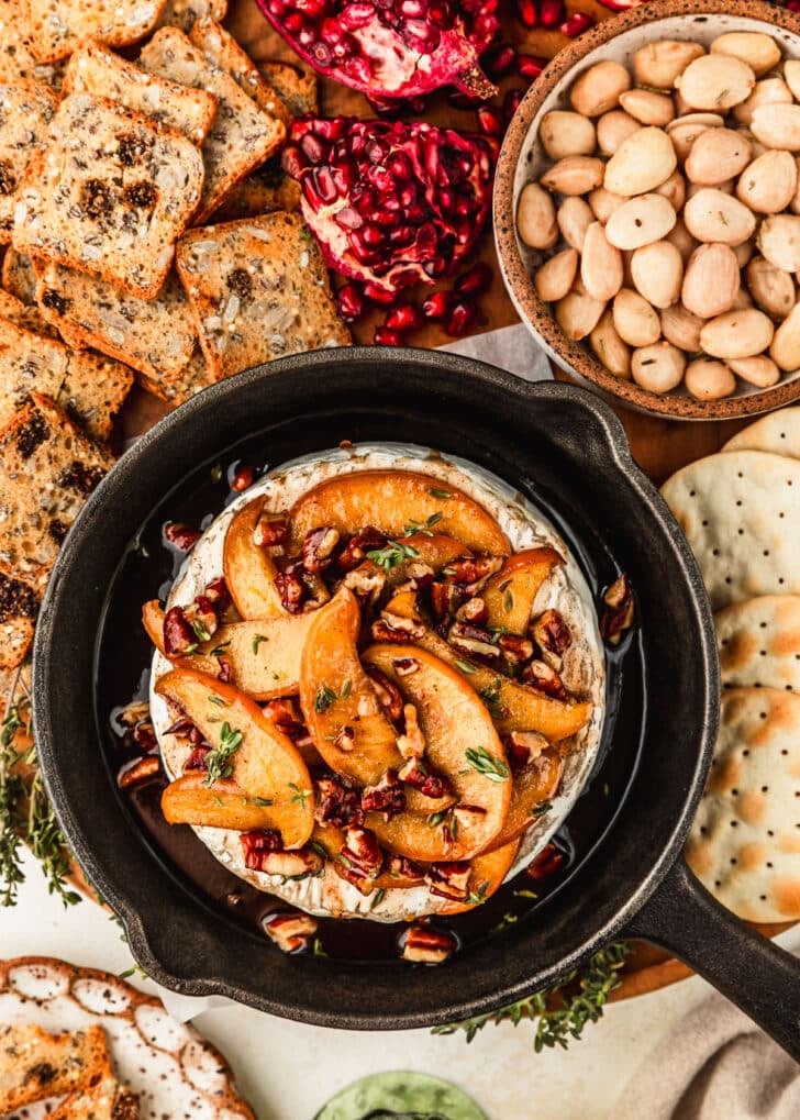 A cast iron skillet of apple baked brie on a wood board next to crackers, nuts, and pomegranates.