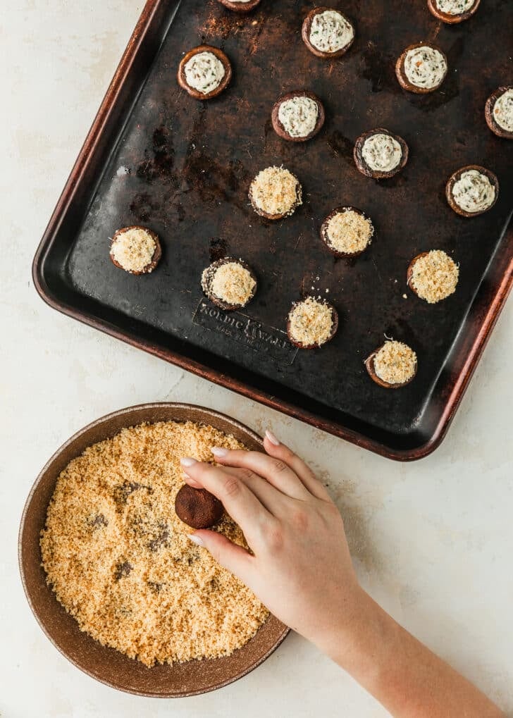 A hand dipping boursin cheese stuffed mushrooms into a brown bowl of Panko breadcrumbs.