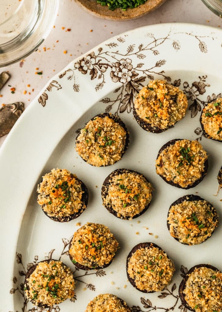 A white and brown floral platter of boursin cheese stuffed mushrooms on a tan counter next to glasses of white wine and a tan bowl of chives.