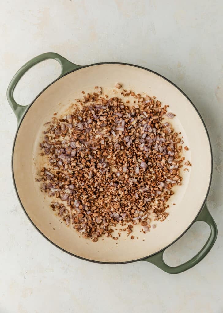 A white skillet with chopped mushrooms and shallots on a beige table.