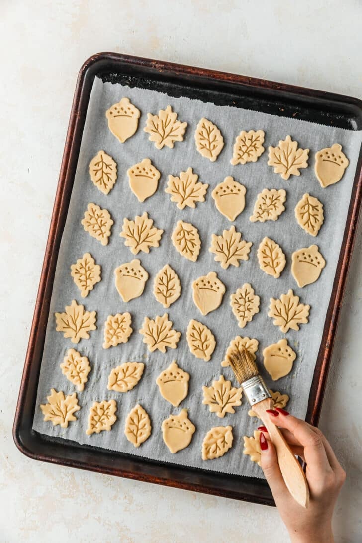 A hand using a pastry brush to brush pie crust leaves with egg wash.
