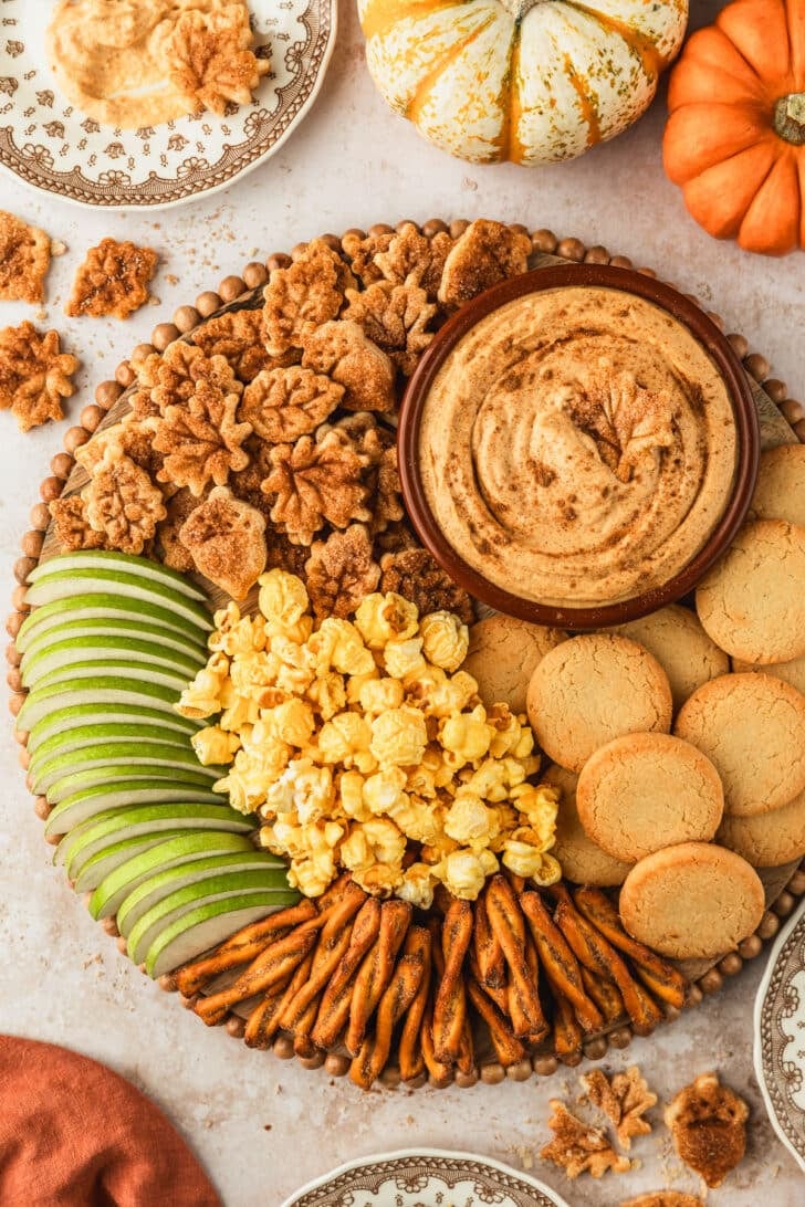 A wood board with a brown bowl of pumpkin pie dip, green apples, cookies, and popcorn on a tan counter next to an orange linen, brown vintage plates, and pumpkins.