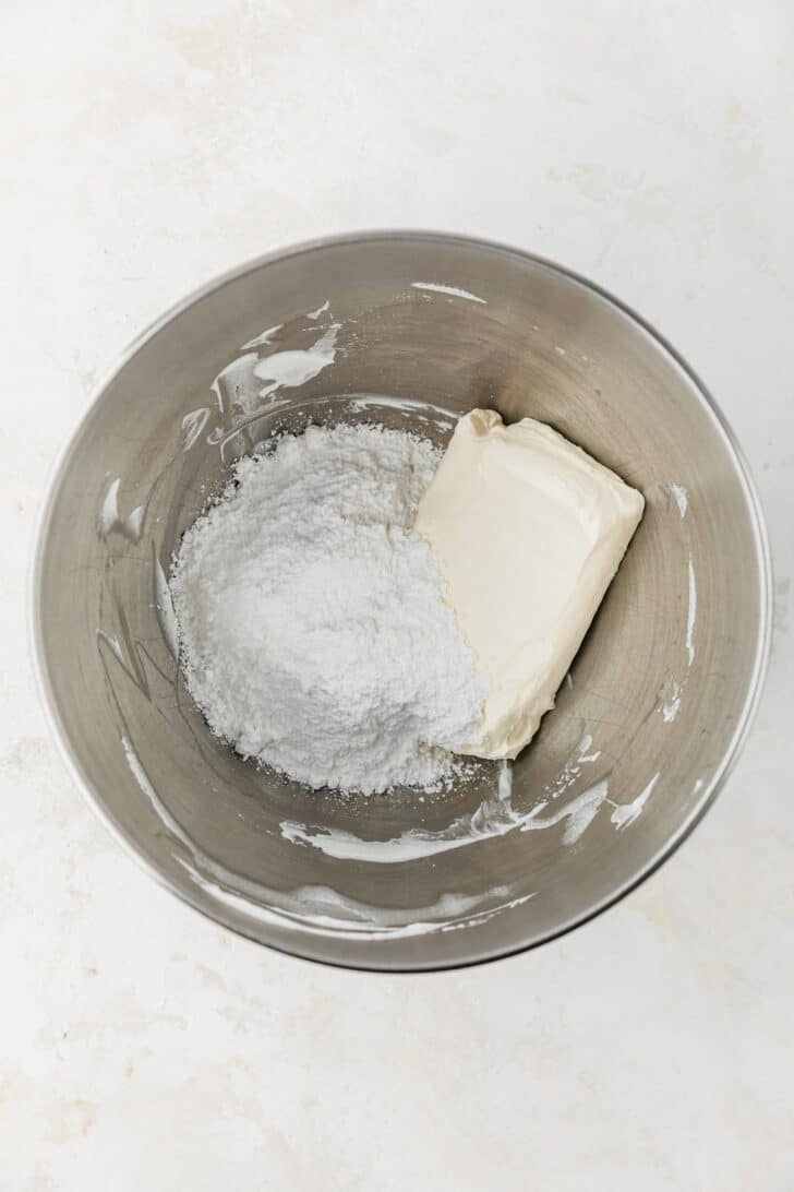 A silver mixing bowl of powdered sugar and cream cheese on a beige table.