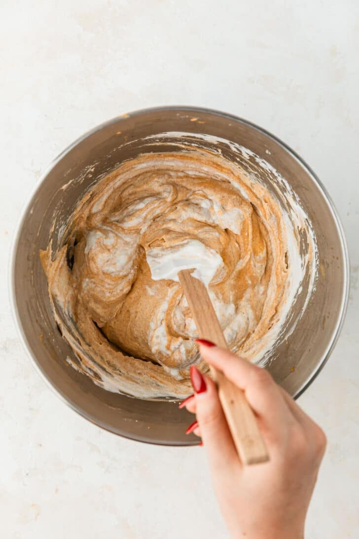 A hand using a rubber spatula to fold whipped cream into pumpkin pie dip in a silver bowl.