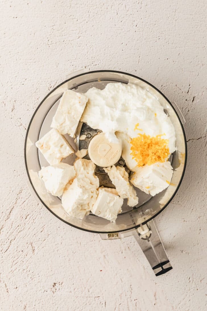 A food processor with cheese, yogurt, and lemon zest on a tan counter.