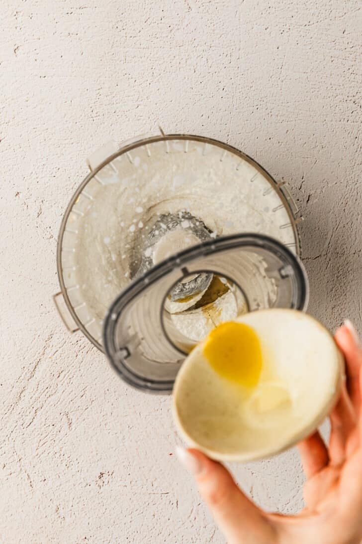 A hand drizzling olive oil into a food processor of cheese dip.