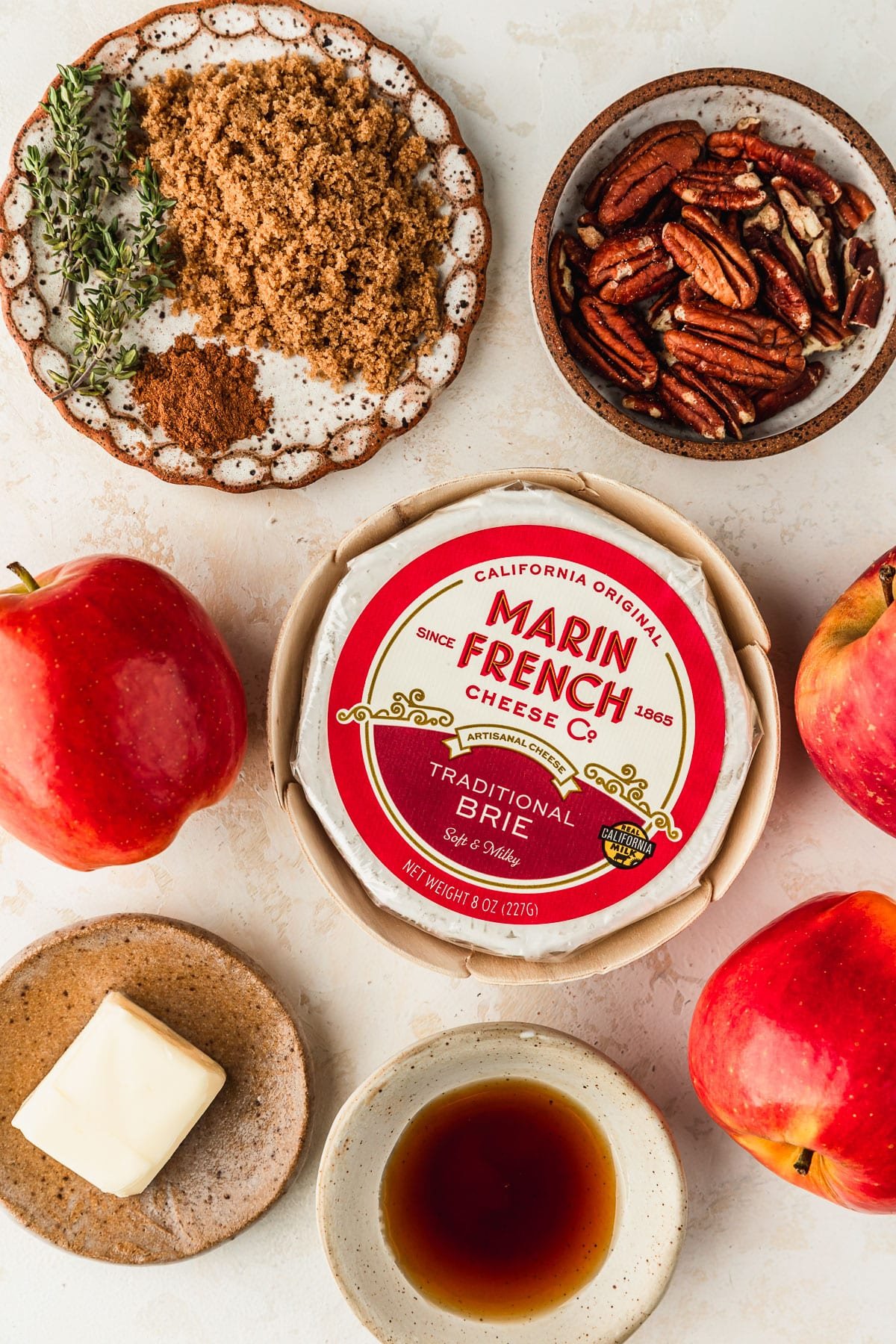 A wheel of brie and apples next to brown and white bowls of pecans, maple syrup, butter, brown sugar, cinnamon, and thyme on a tan table.