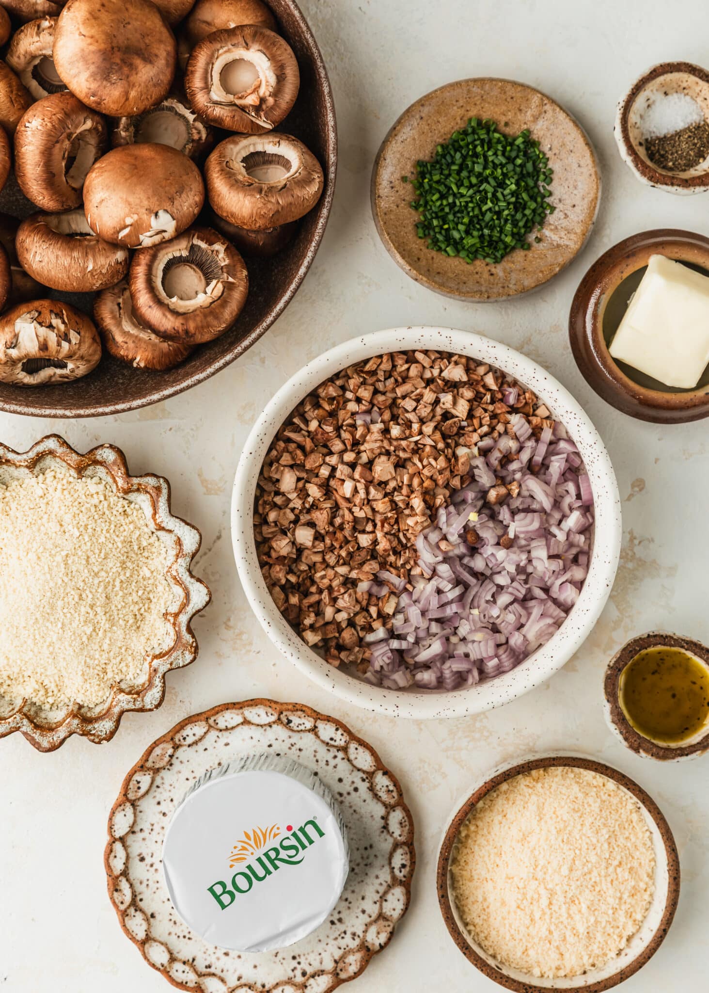 Brown and white bowls of mushrooms, shallots, parmesan, chives, spices, butter, oil, Panko, and cheese on a tan counter.