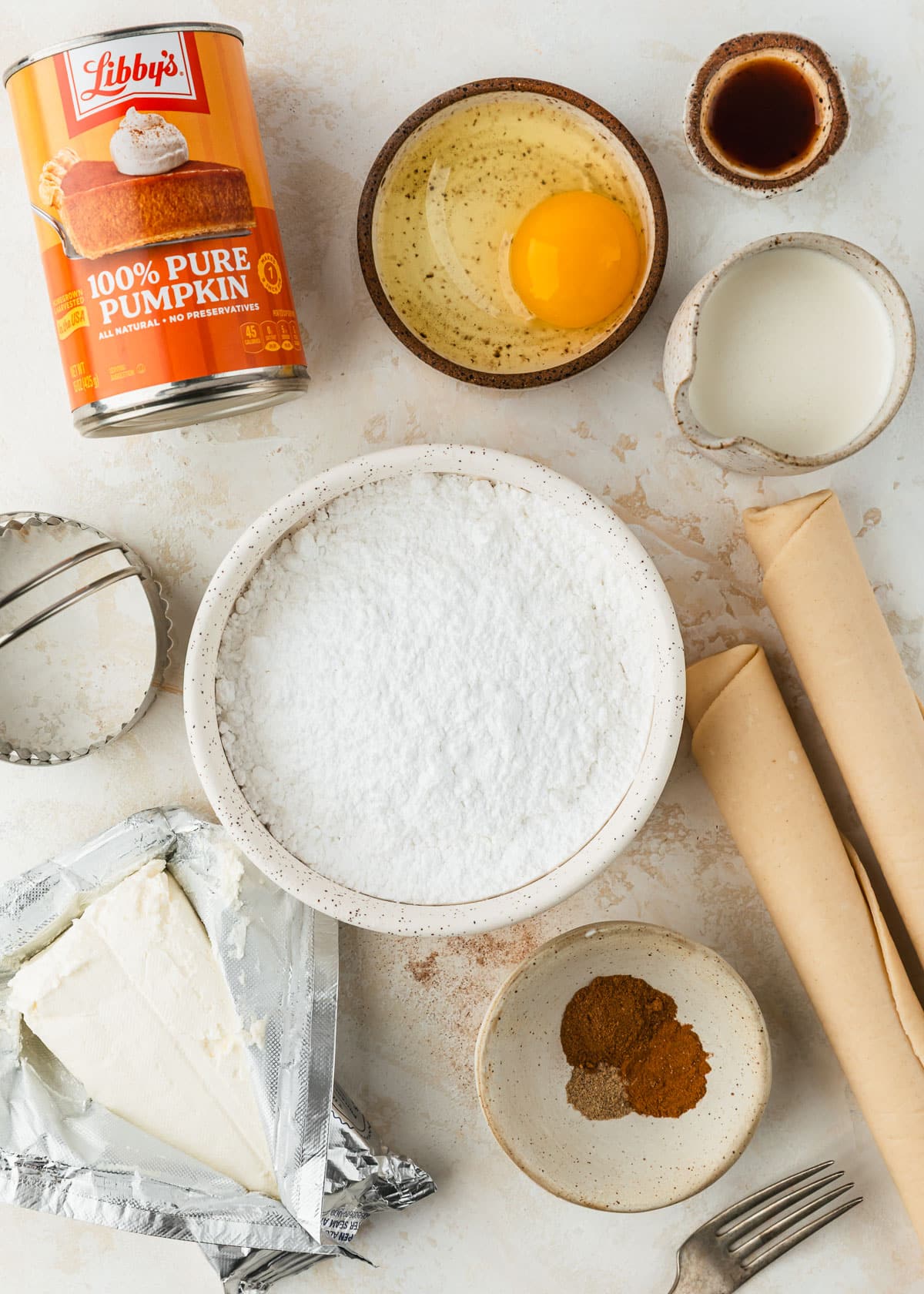 White and brown bowls of powdered sugar, an egg, cream, vanilla, and spices next to pie dough, cream cheese, and a can of pumpkin on a tan counter.