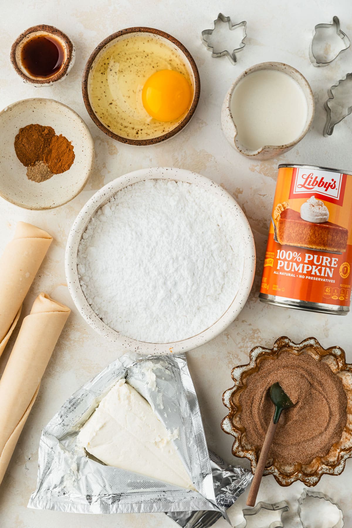White and brown bowls of powdered sugar, cinnamon sugar, an egg, spices, vanilla, and cream next to pie crust, a block of cream cheese, and a can of pumpkin on a tan counter.