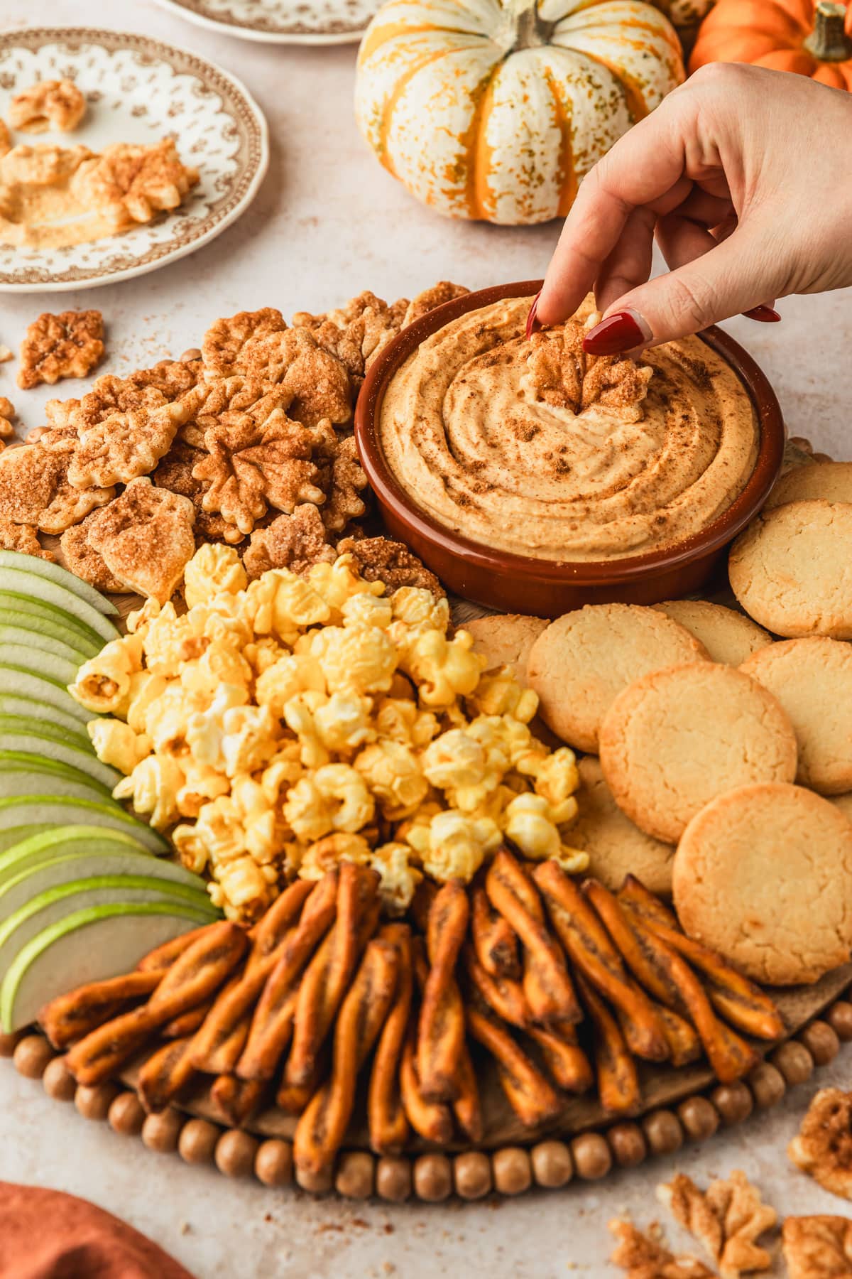 A hand dipping a cookie into a brown bowl of pumpkin pie dip on a wood board of cookies, popcorn, and green apples next to vintage brown plates and pumpkins with a tan stone background.