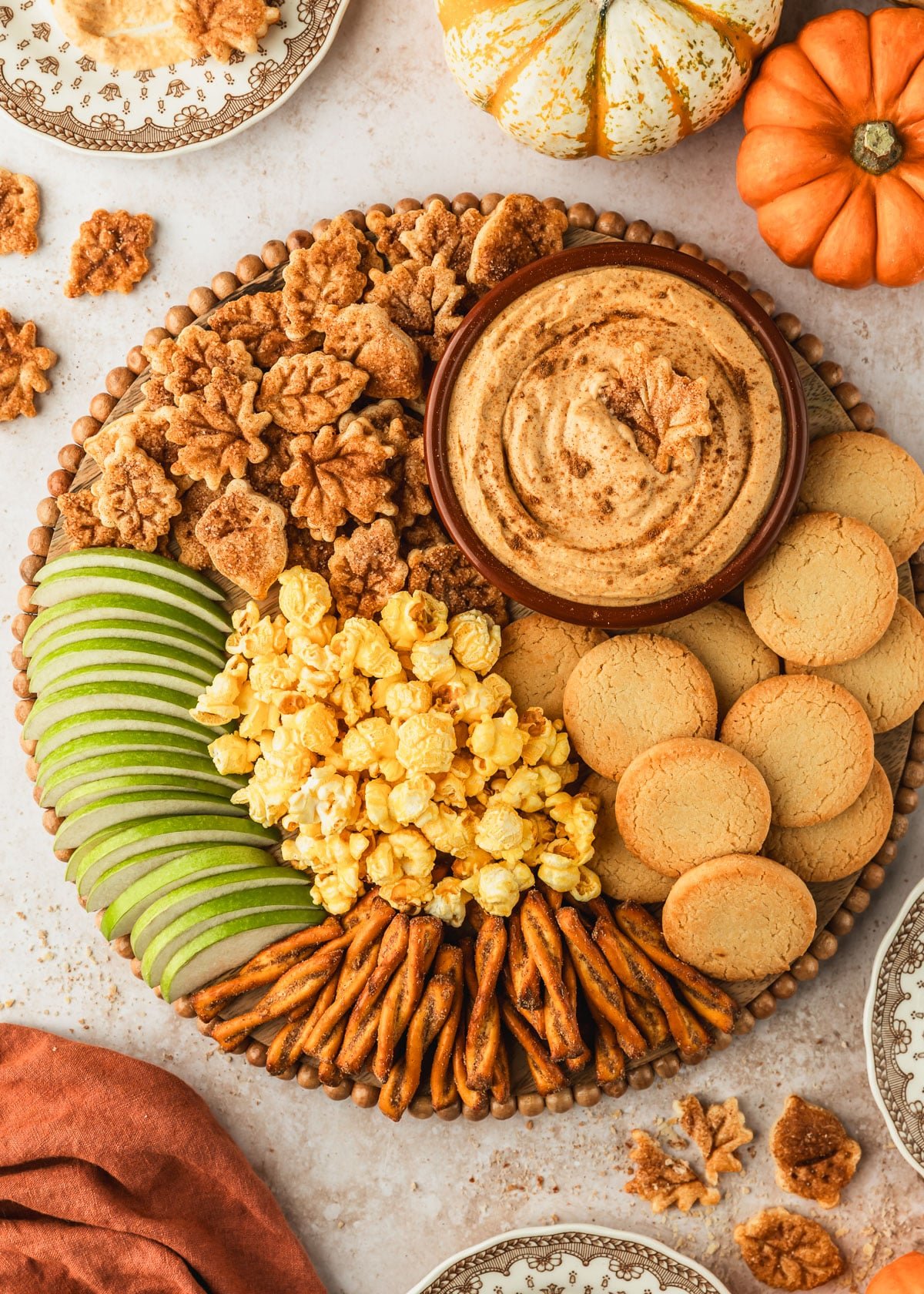 A wood tray with a brown bowl of pumpkin pie dip, green apple slices, leaf-shaped cookies, and popcorn on a tan counter next to an orange linen, vintage brown and white plates, and pumpkins.